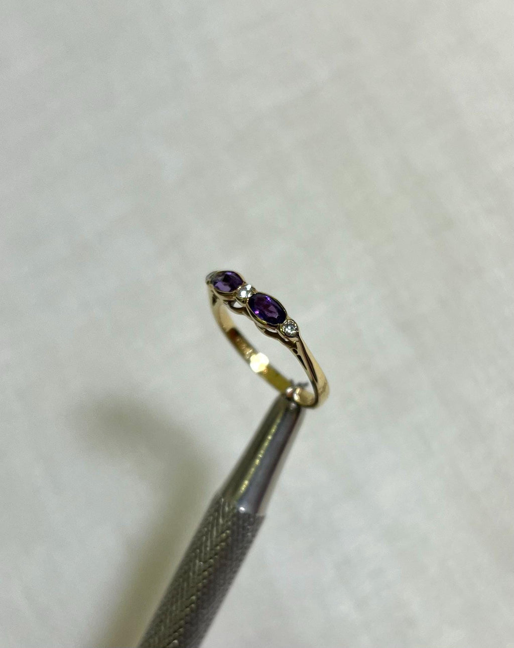 Gold ring with purple gemstone held by tweezers on a light beige background