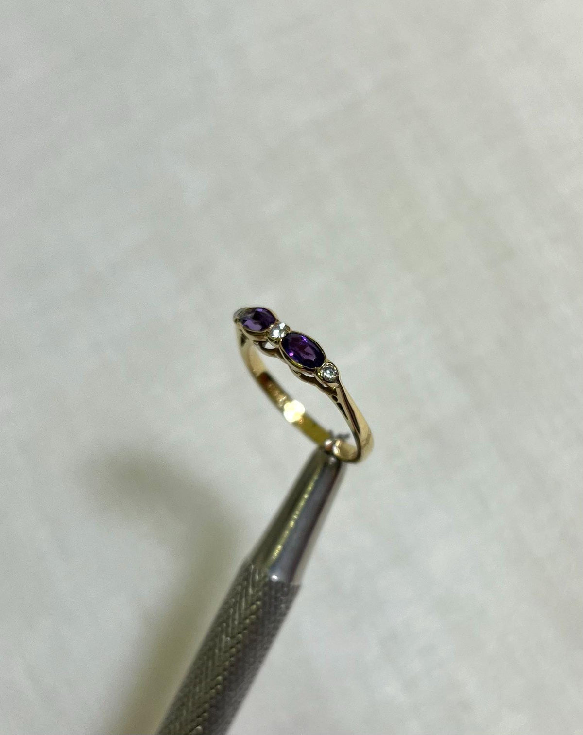Gold ring with purple gemstone held by tweezers on a light beige background
