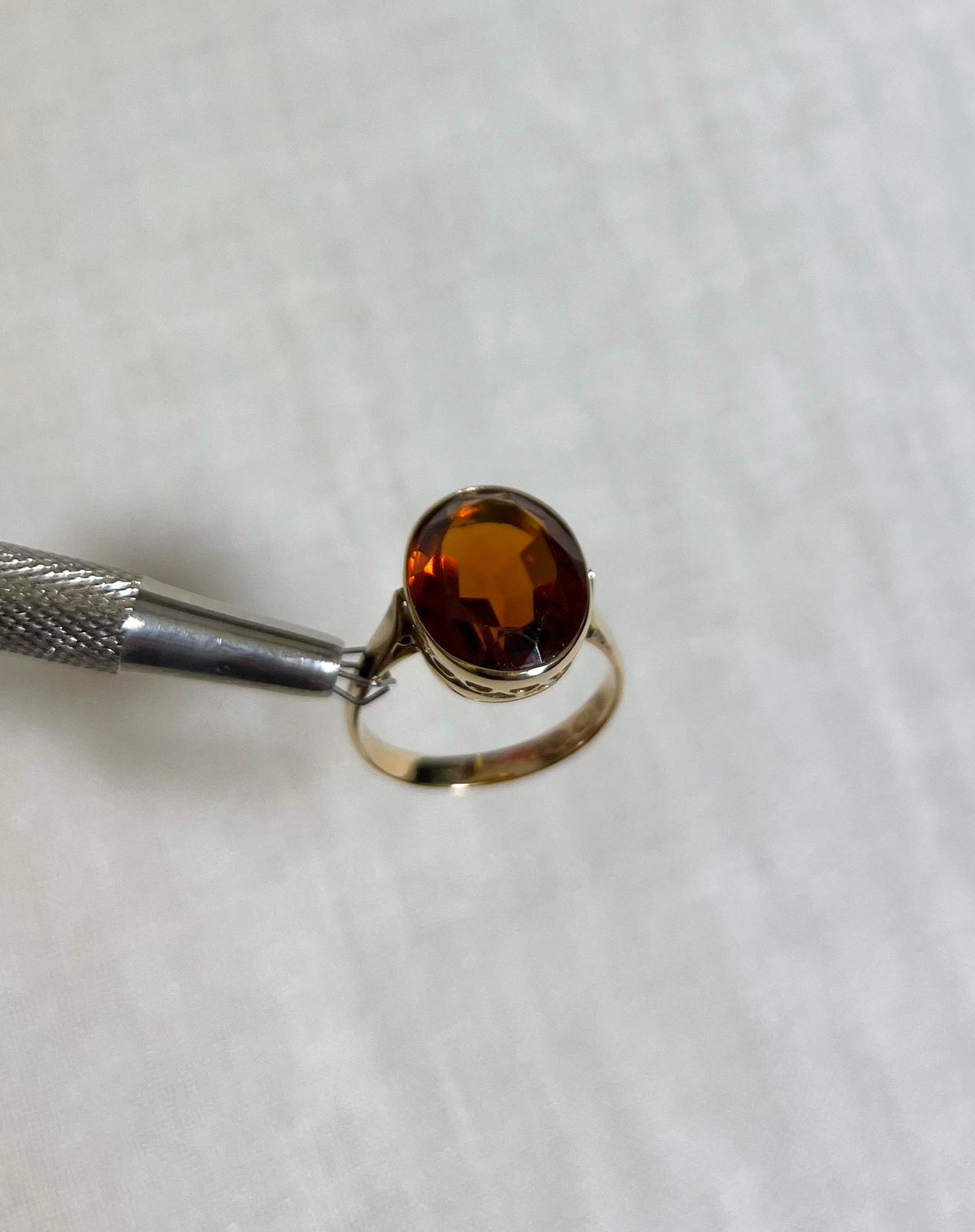 Gold ring with oval citrine stone held by silver tool against white fabric background 