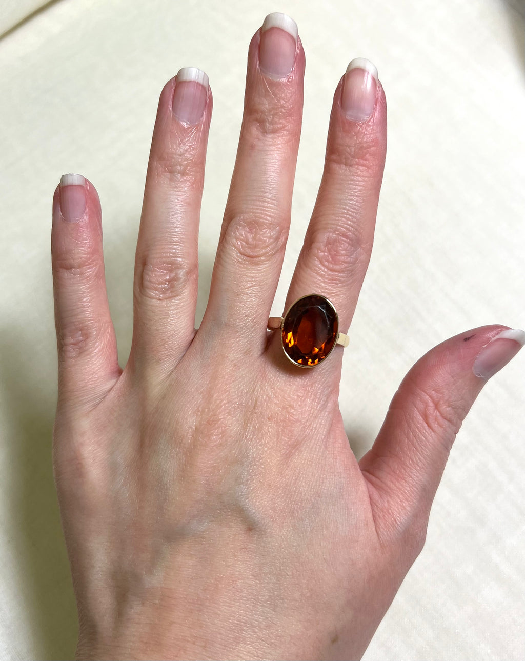A gold ring with an oval citrine  gemstone worn on a person's finger, shown against a light beige fabric background.