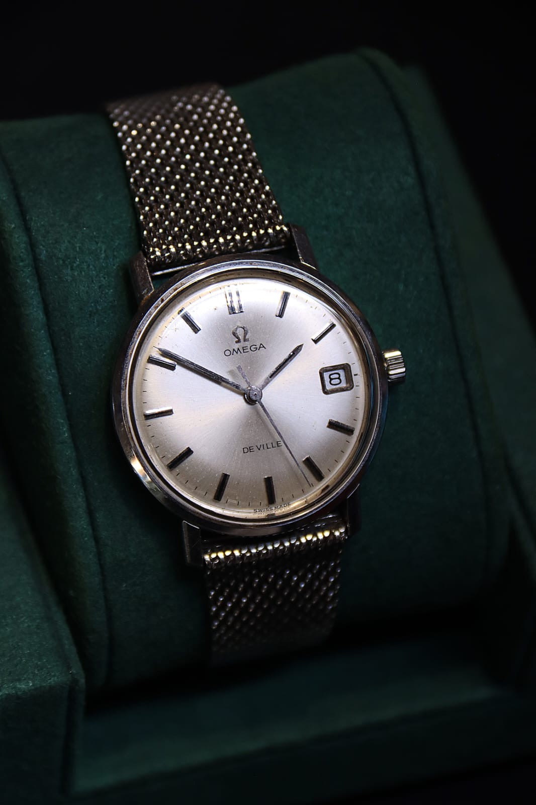 A vintage Omega De Ville wristwatch with a silver dial and stainless steel mesh strap, displayed on dark green velvet watch cushion.