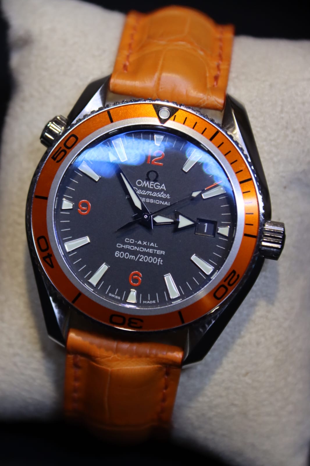 Omega Seamaster Planet Ocean watch with black dial, orange bezel and orange strap, resting on white cushion