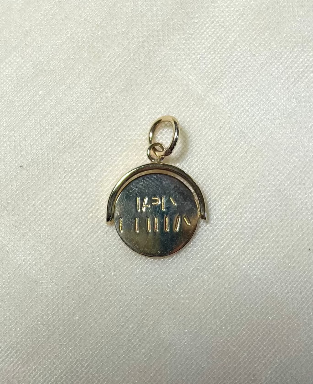 A small round gold pendant charm with a bail at the top and engraved happy birthday message, photographed on a light fabric background. 