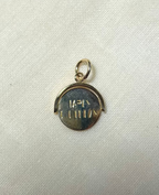 A small round gold pendant charm with a bail at the top and engraved happy birthday message, photographed on a light fabric background. 