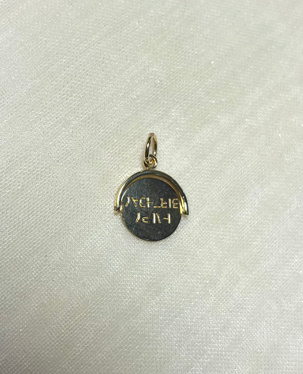 A small round gold pendant charm with engraved text, displayed on a textured off-white fabric background.