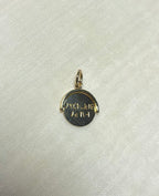 A small round gold pendant charm with engraved text, displayed on a textured off-white fabric background.