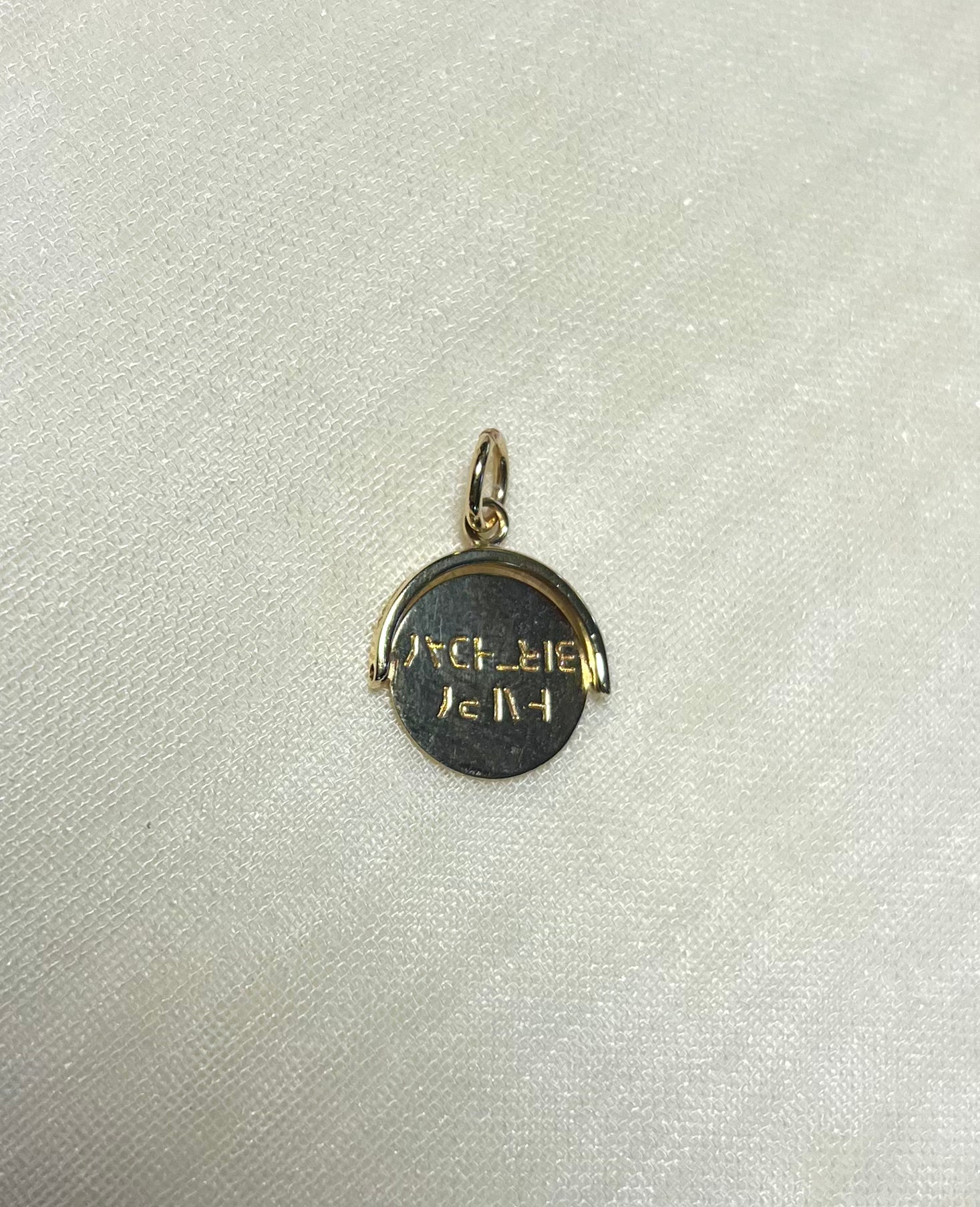 A small round gold pendant charm with engraved text, displayed on a textured off-white fabric background.