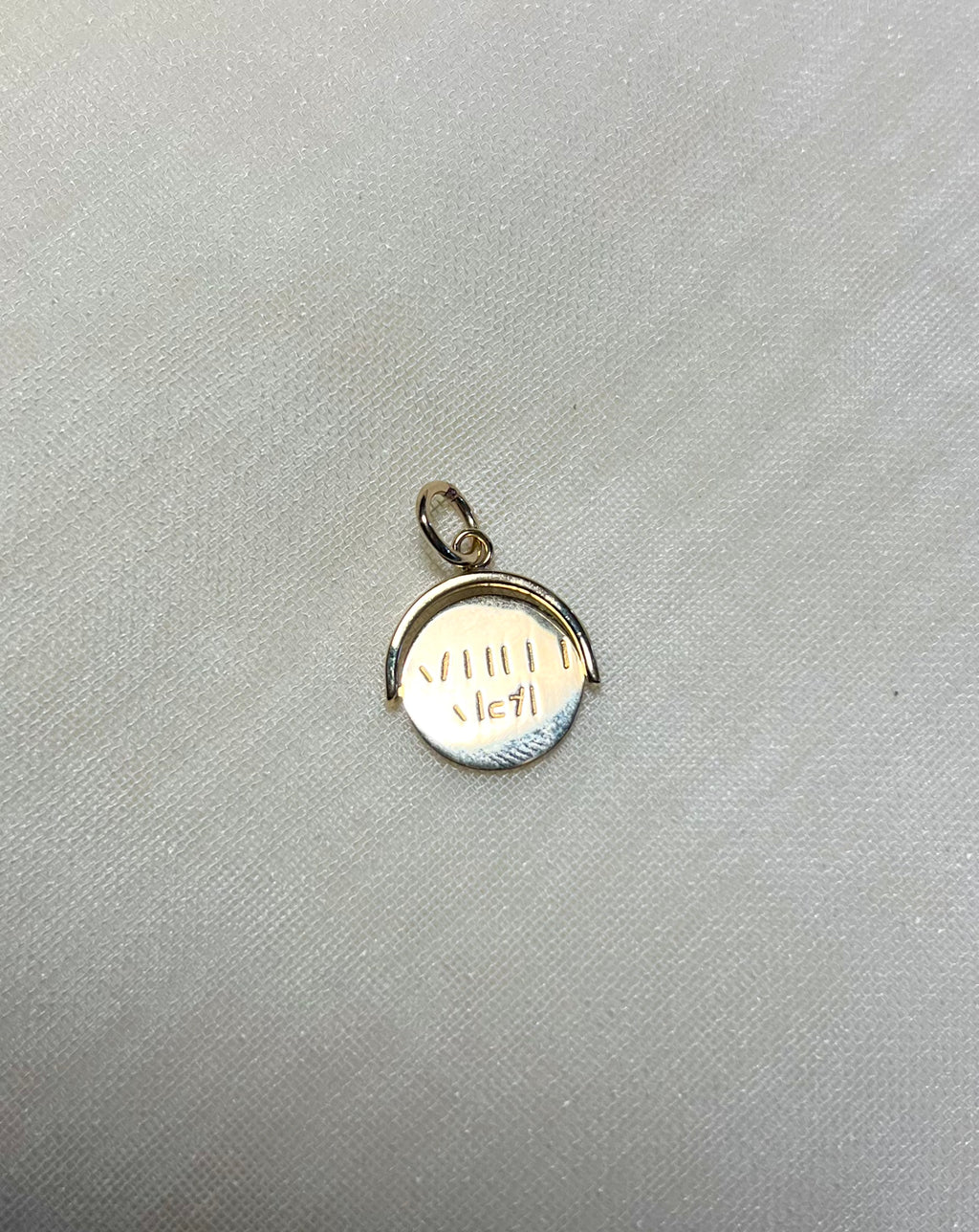 A small gold pendant charm with a circular design and a bail at the top, resting on a textured light gray fabric background.