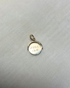 A small gold pendant charm with a circular design and a bail at the top, resting on a textured light gray fabric background.