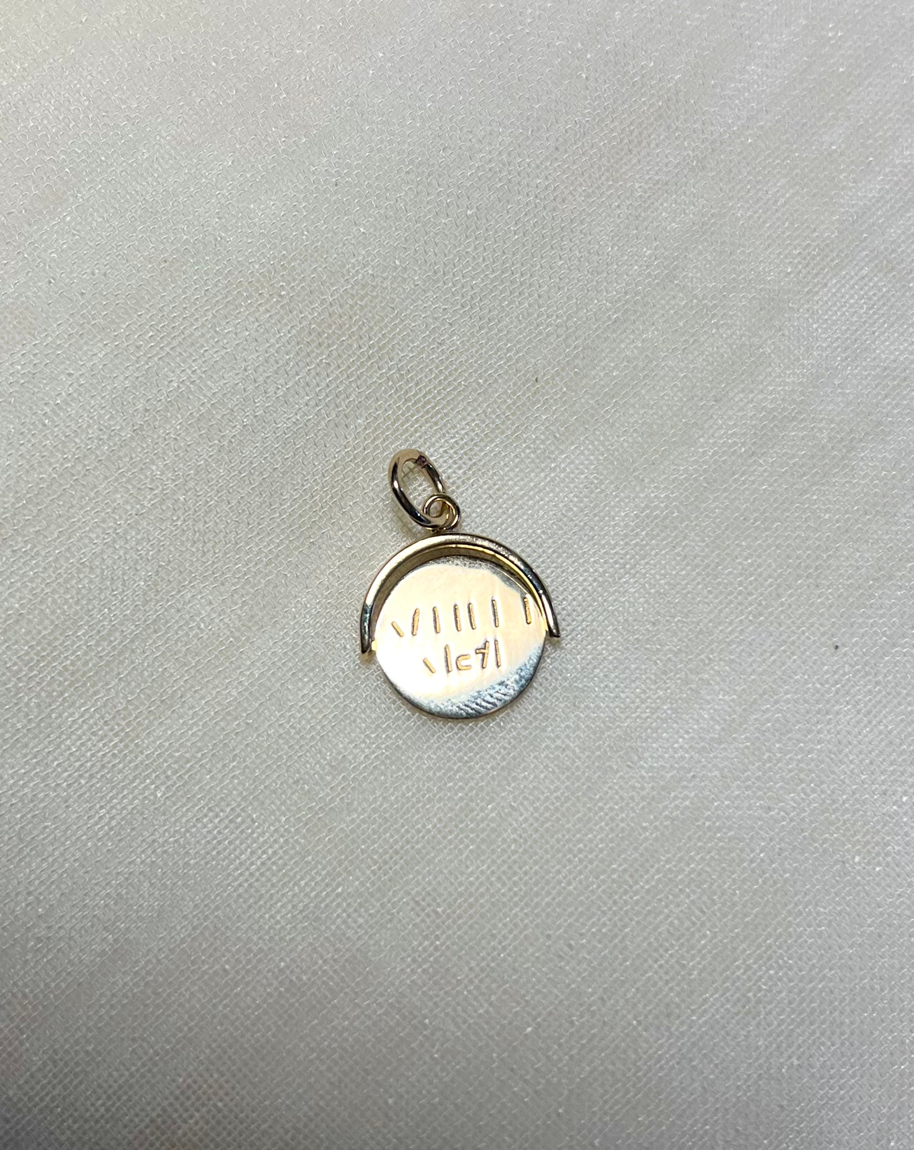 A small gold pendant charm with a circular design and a bail at the top, resting on a textured light gray fabric background.
