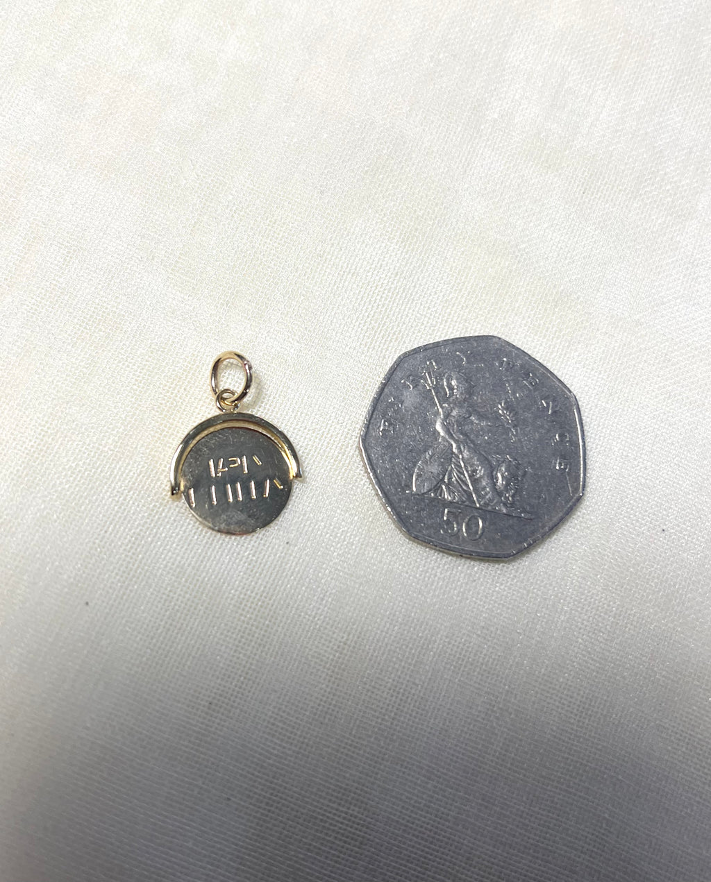 A small round gold pendant with happy birthday engraving, shown next to a silver 50 pence coin for scale on a light fabric background.