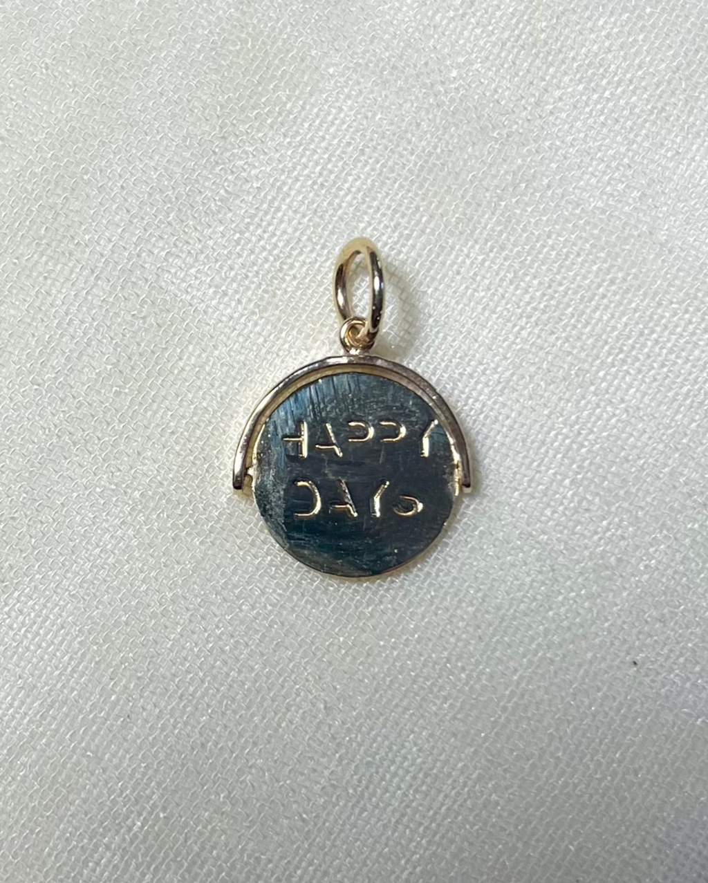 a gold round charm with 'happy days' engraved, resting on cream linen background