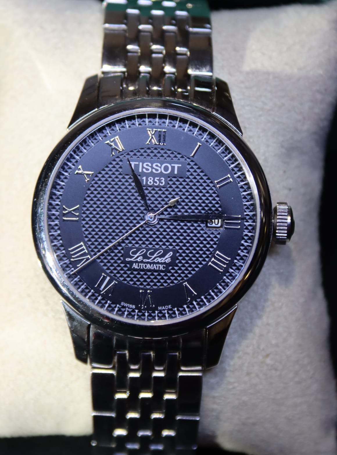 A Tissot Le Locle Automatic watch with black dial on stainless steel bracelet, displayed on cream cushion
