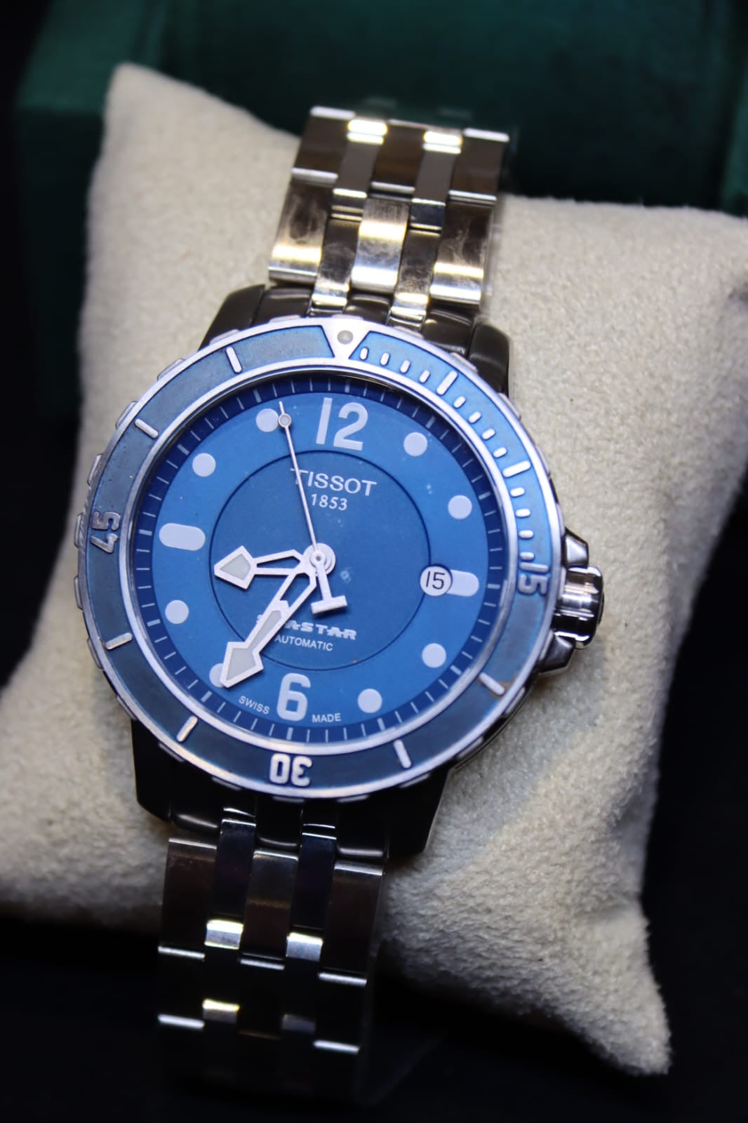 A Tissot Seastar automatic watch with a blue dial and blue bezel, paired with a silver stainless steel link bracelet, displayed on a beige cushion.