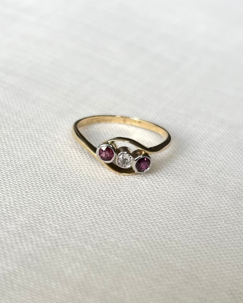 A gold ring with three small gemstones in the center, featuring two pink round rubies stones and centre diamond round stone, displayed on a textured white fabric background.