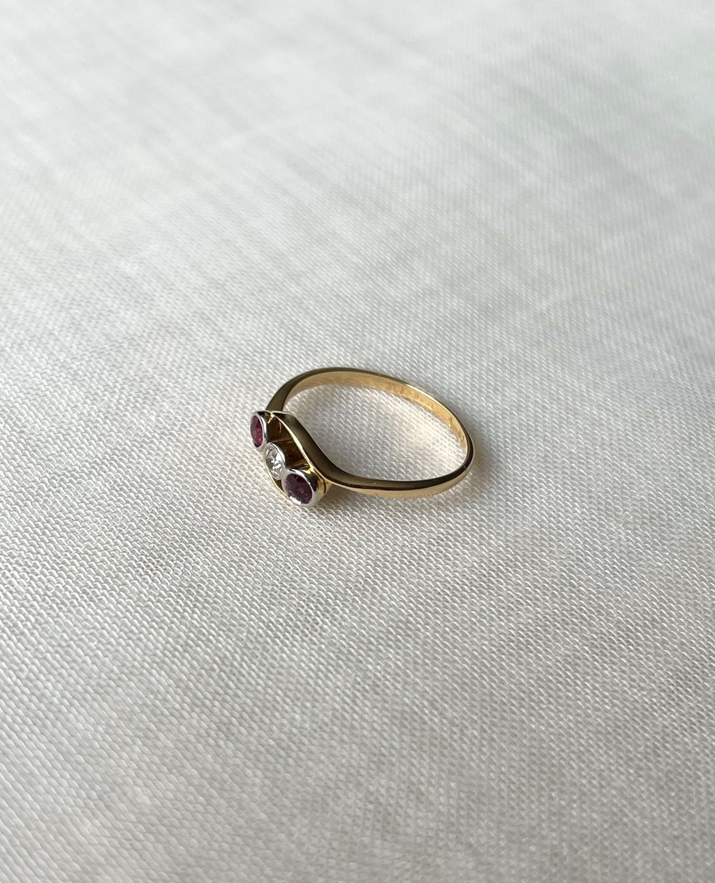 Side view of a gold ring with three small gemstones (two red, one clear) resting on a textured white linen surface.