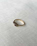 Side view of a gold ring with three small gemstones (two red, one clear) resting on a textured white linen surface.