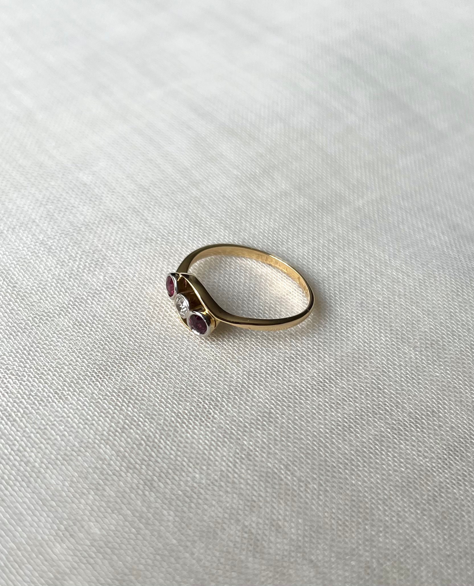 Side view of a gold ring with three small gemstones (two red, one clear) resting on a textured white linen surface.