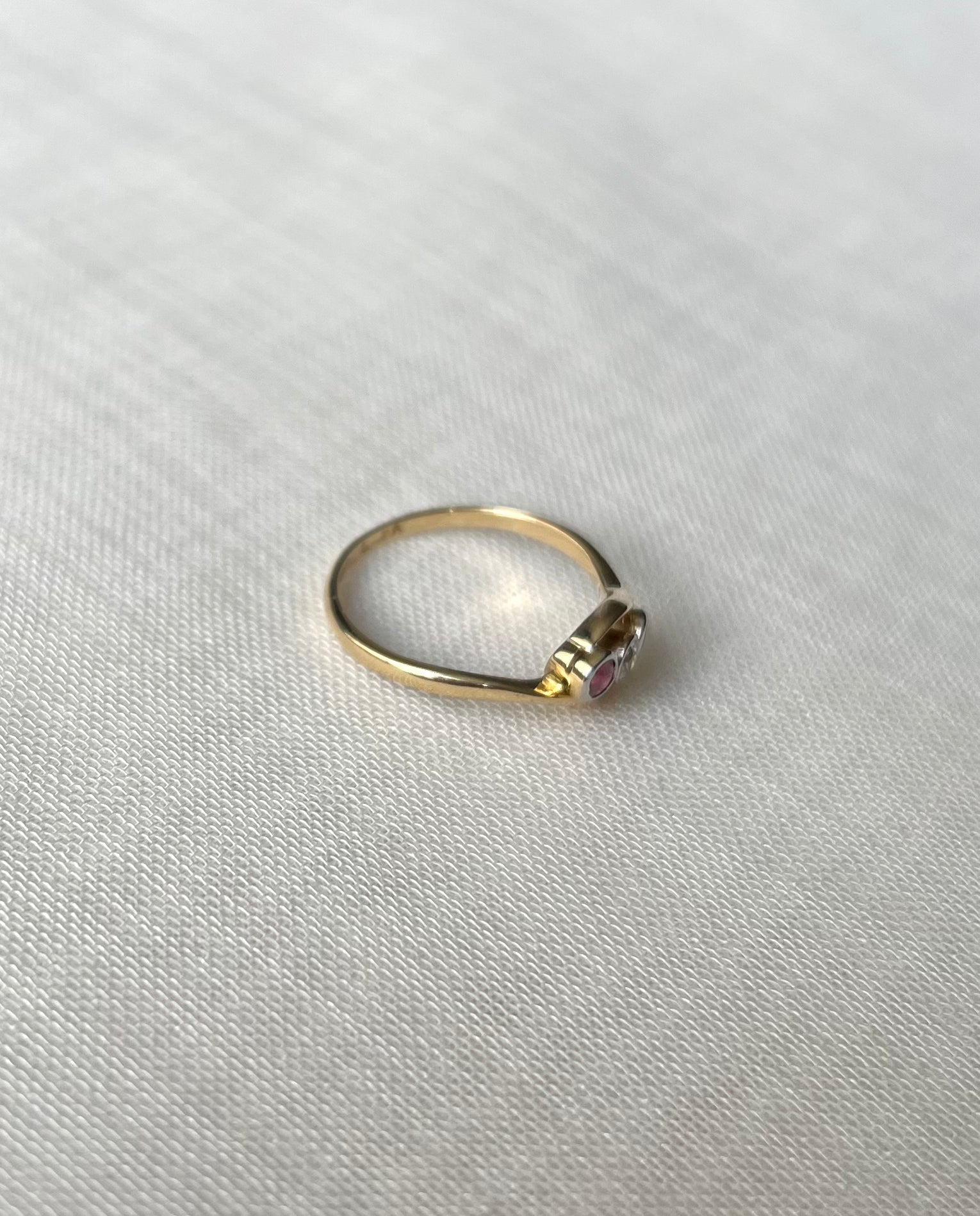 side view of a delicate gold ring with three round gemstones - 2 pink, one clear, resting on a textured white fabric background.