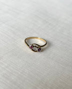 A gold ring with three small stones in the center, featuring two dark pink rubies and one clear round diamond  stone, displayed on a textured white fabric background.