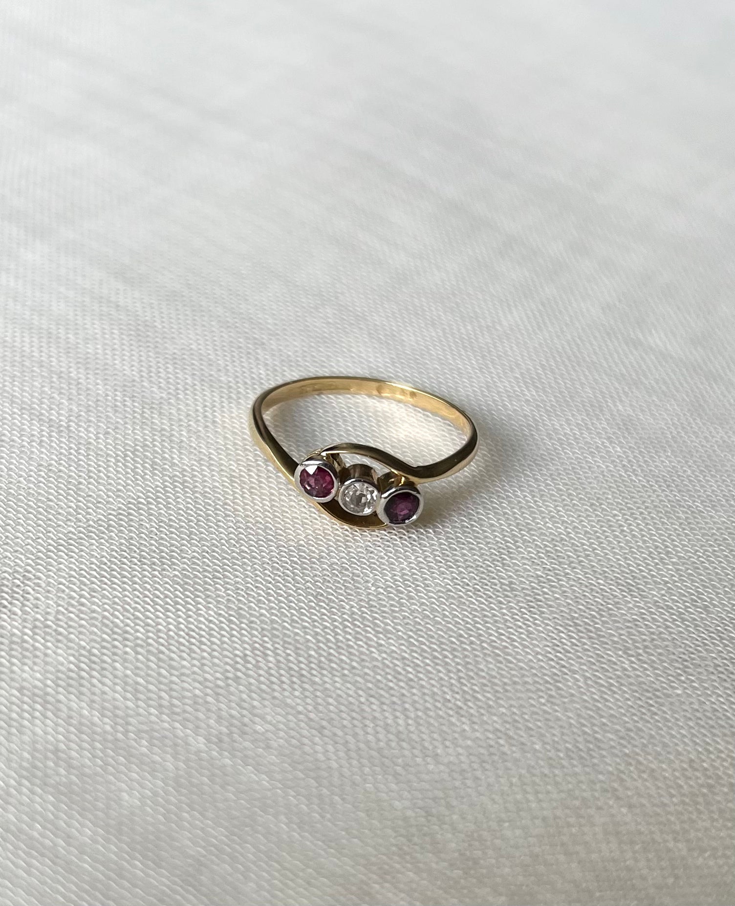 A gold ring with three small stones in the center, featuring two dark pink rubies and one clear round diamond  stone, displayed on a textured white fabric background.