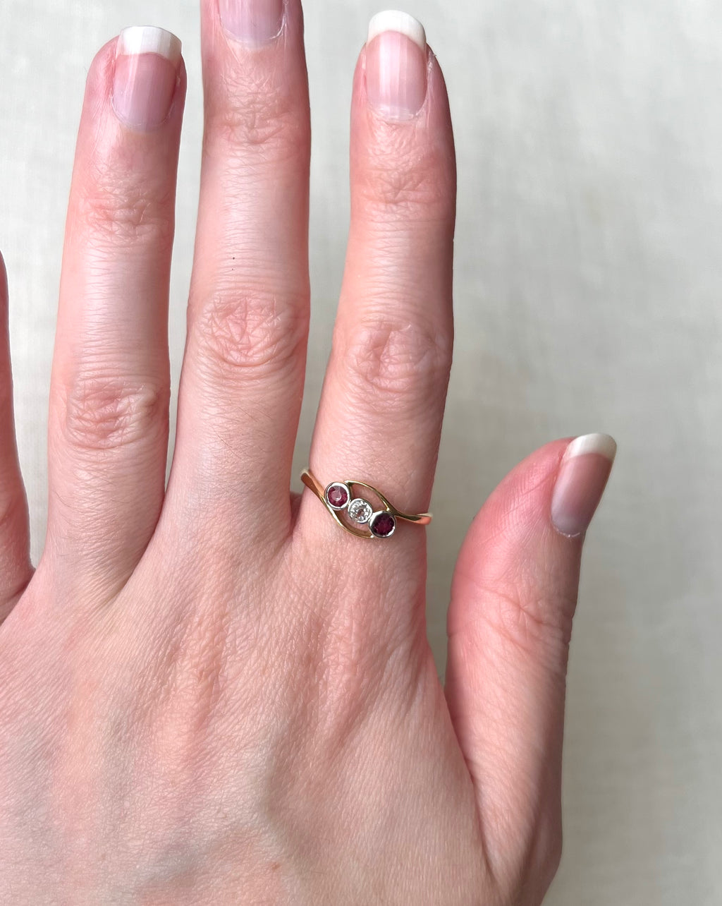 A gold ring with three small round gemstones of rubies and a diamond with crossover design, worn on a person's finger against a light neutral background.