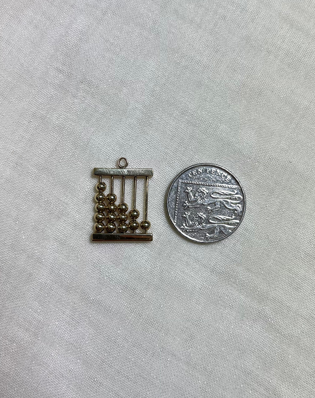 gold abacus charm next to ten pence coin for scale on white fabric background