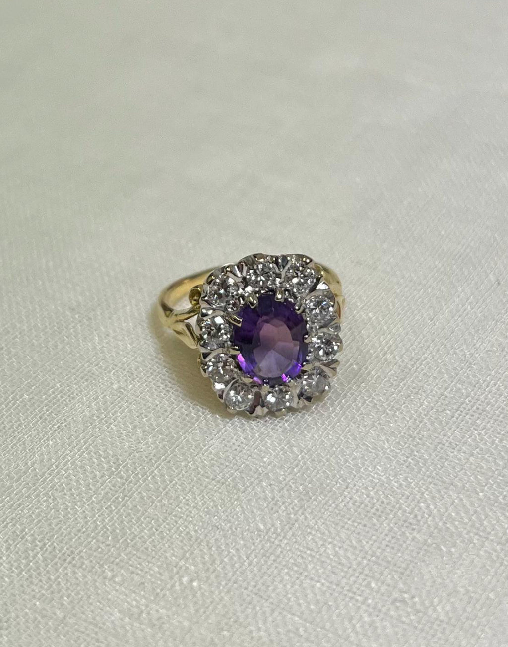 gold ring with purple centre stone surrounded by sparkling diamonds, resting on white fabric background