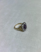 gold ring with purple centre gemstone surrounded by sparkling diamonds, resting on white fabric background