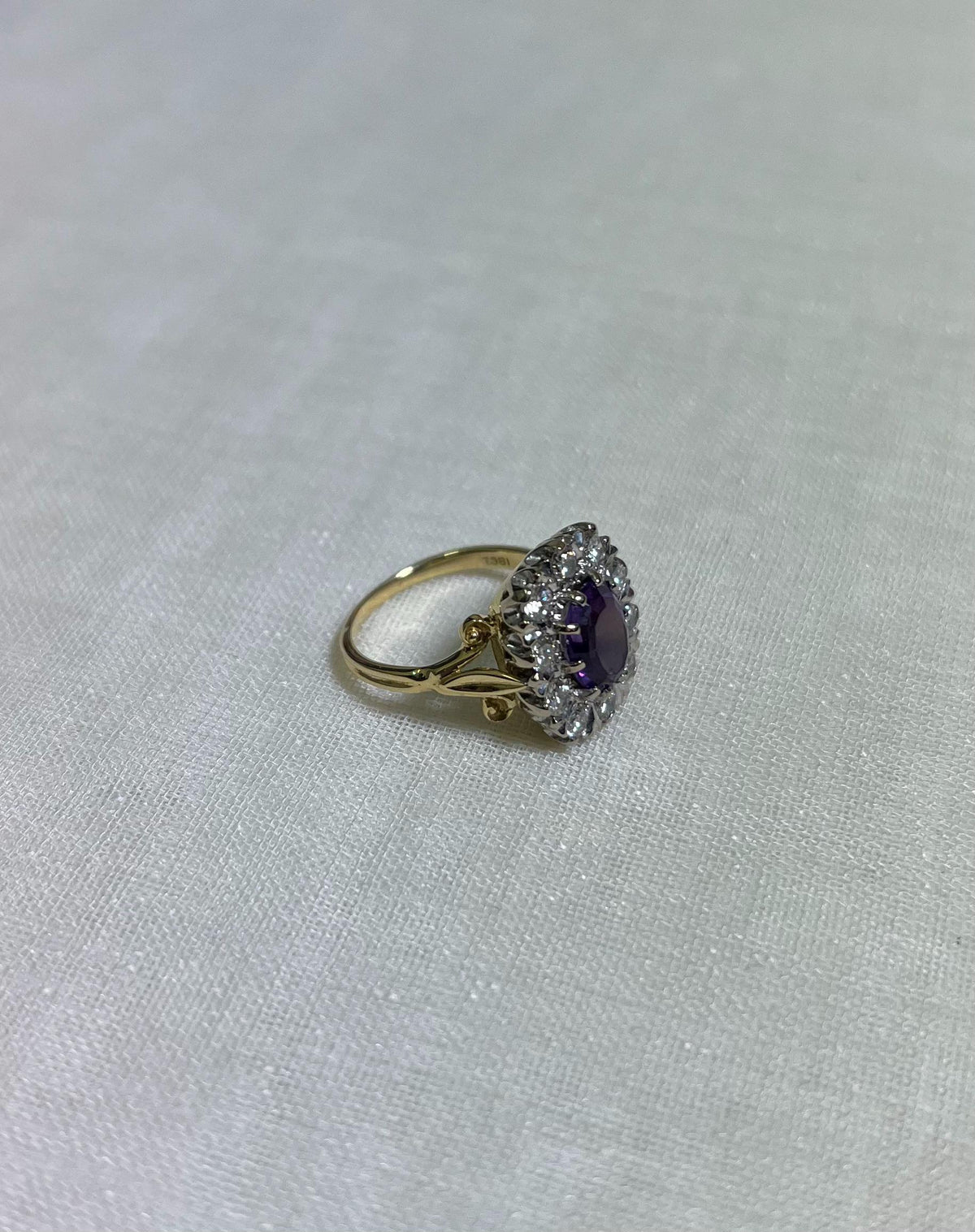 gold ring with purple centre gemstone surrounded by sparkling diamonds, resting on white fabric background