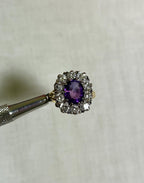 gold ring with purple centre gemstone surrounded by sparkling diamonds, held by silver tool above white background 