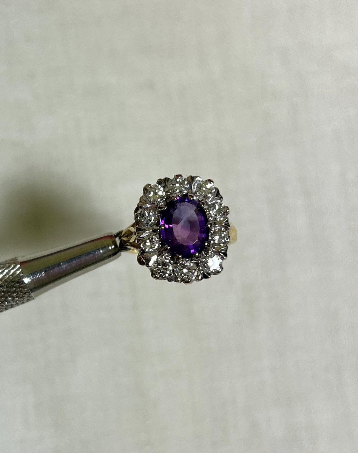 gold ring with purple centre gemstone surrounded by sparkling diamonds, held by silver tool above white background 