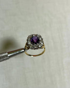 gold ring with purple gemstone surrounded by clear gemstone held by silver metal tool above white background 