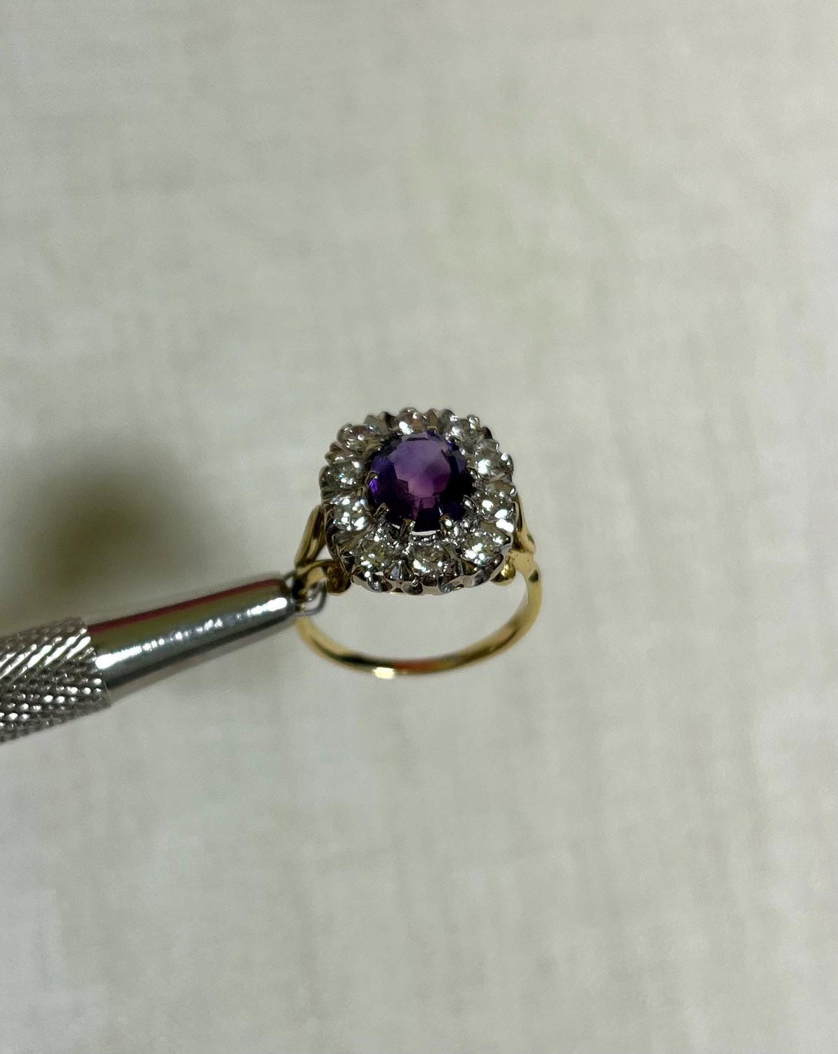 gold ring with purple gemstone surrounded by clear gemstone held by silver metal tool above white background 