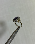 side view of gold ring with purple gemstone and diamonds, held by silver tool above white background