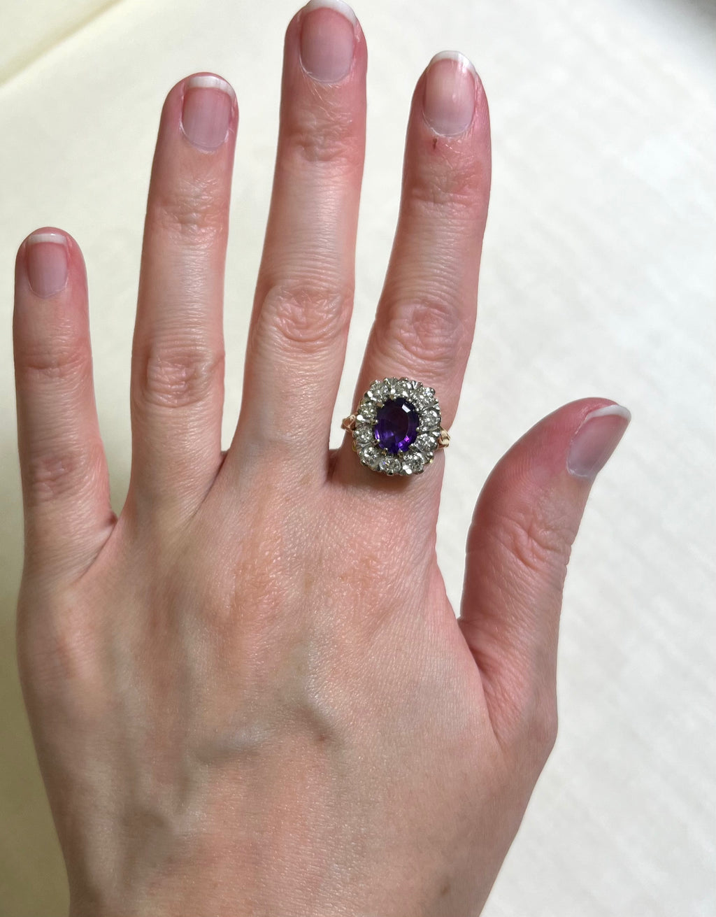 gold ring with purple gemstone surrounded by diamonds, worn on finger, white background