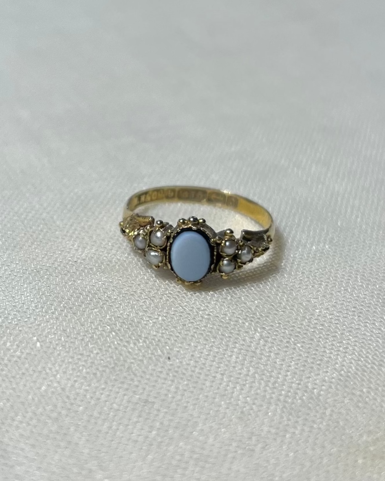 an antique gold ring with a light blue oval sardonyx centre stone with seed pearls either side, resting on a cream linen background 
