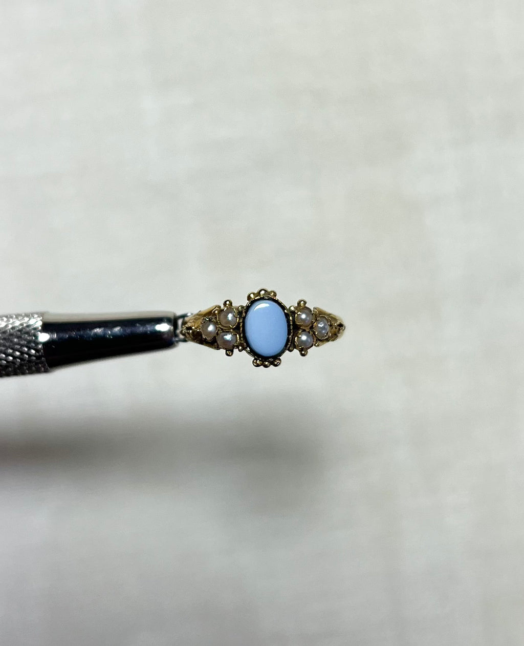 an antique gold ring set with seed pearl and an oval sardonyx stone in the centre, held by silver tool above cream coloured background 