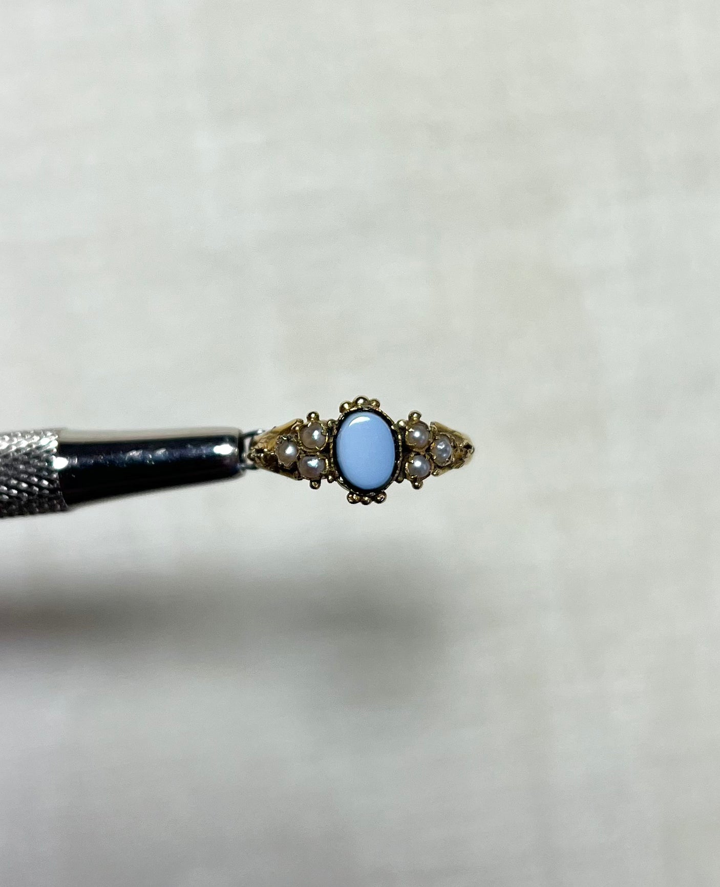 an antique gold ring set with seed pearl and an oval sardonyx stone in the centre, held by silver tool above cream coloured background 