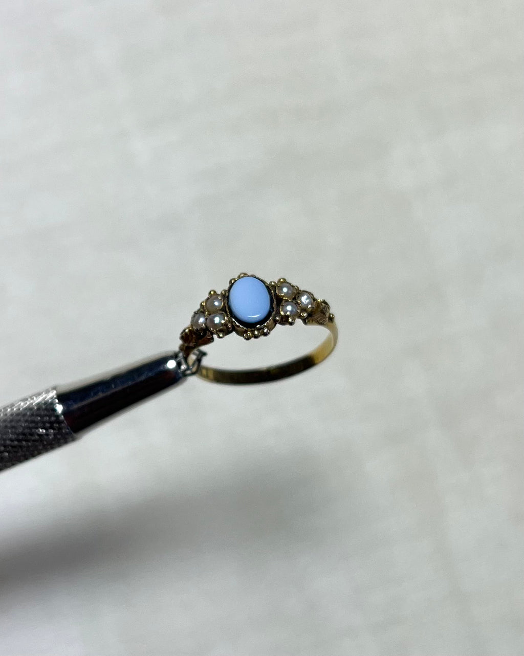 an antique gold ring with light blue oval sardonyx and seed pearls, held by silver tool above cream coloured background 