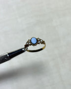 an antique gold ring with light blue oval sardonyx and seed pearls, held by silver tool above cream coloured background 