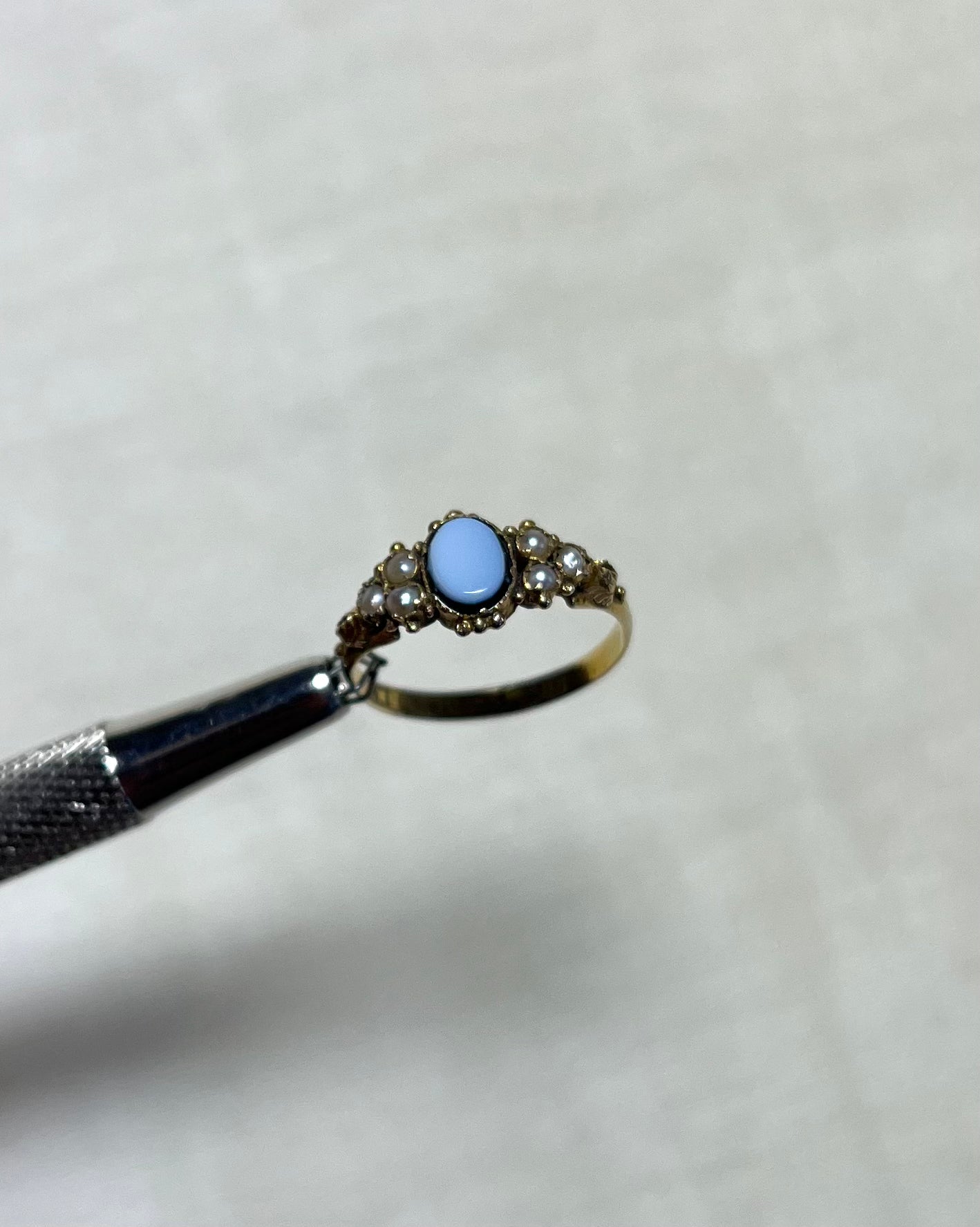 an antique gold ring with light blue oval sardonyx and seed pearls, held by silver tool above cream coloured background 