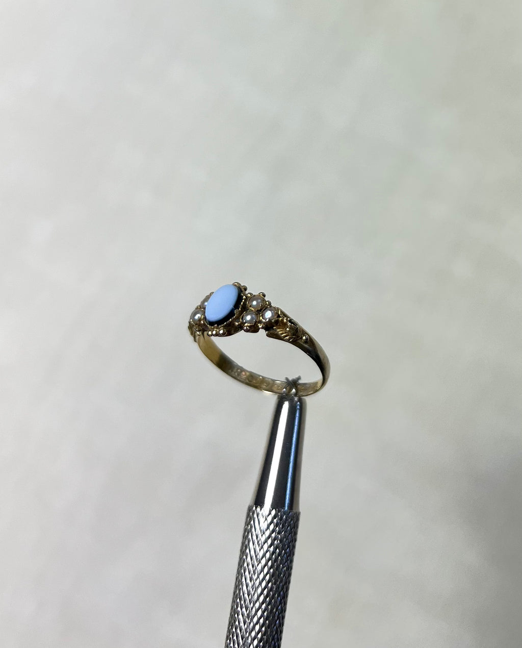 side view of antique gold ring with light blue stone and pearls, help by silver tool above cream coloured background 