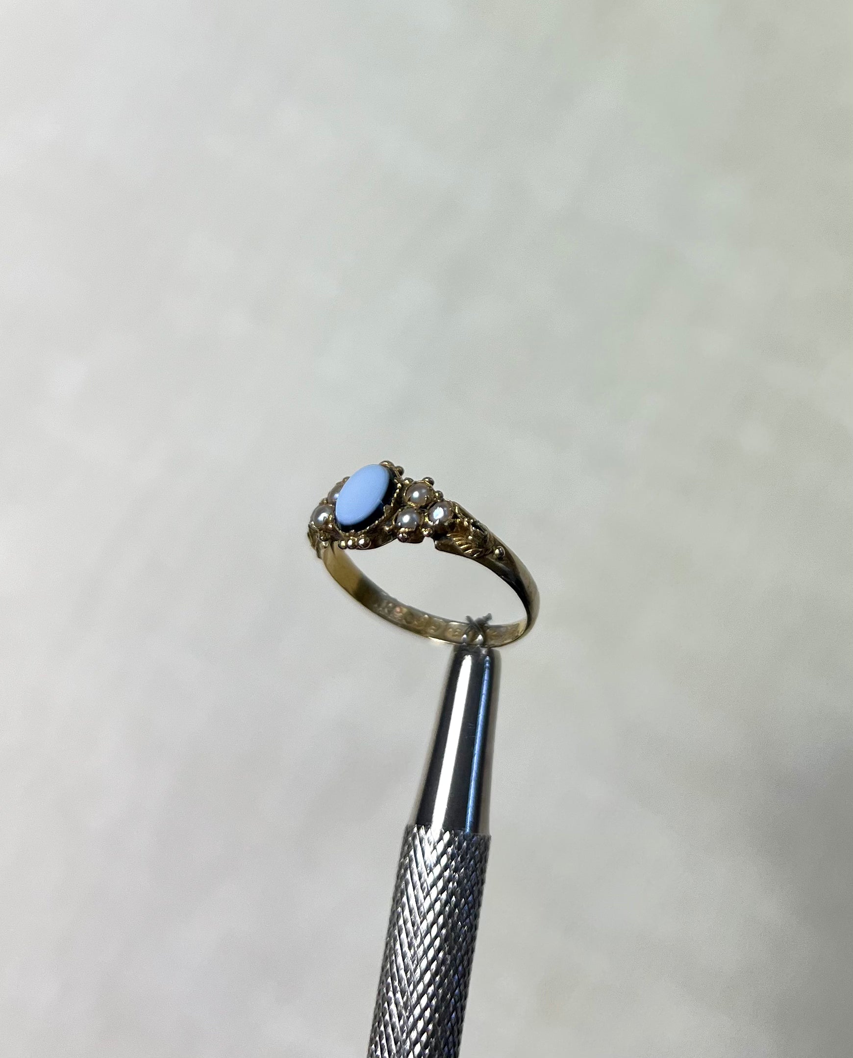 side view of antique gold ring with light blue stone and pearls, help by silver tool above cream coloured background 