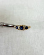 A gold ring with 3 blue stones held by silver tool against white fabric background 