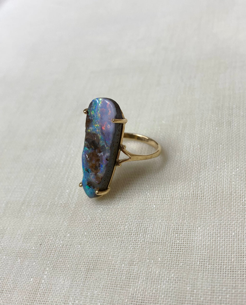 A gold ring featuring a large, irregularly shaped natural opal gemstone with vibrant blue, green, and orange play-of-color, set on a textured beige fabric background.