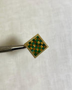 gold square ring with green and brown checkerboard pattern held by silver tool on white fabric background