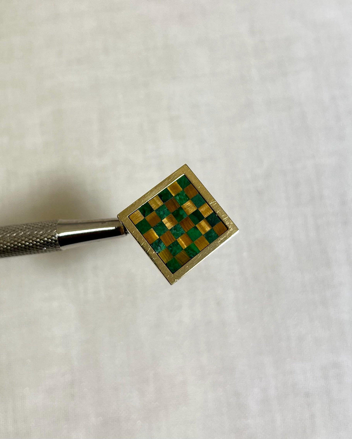 gold square ring with green and brown checkerboard pattern held by silver tool on white fabric background