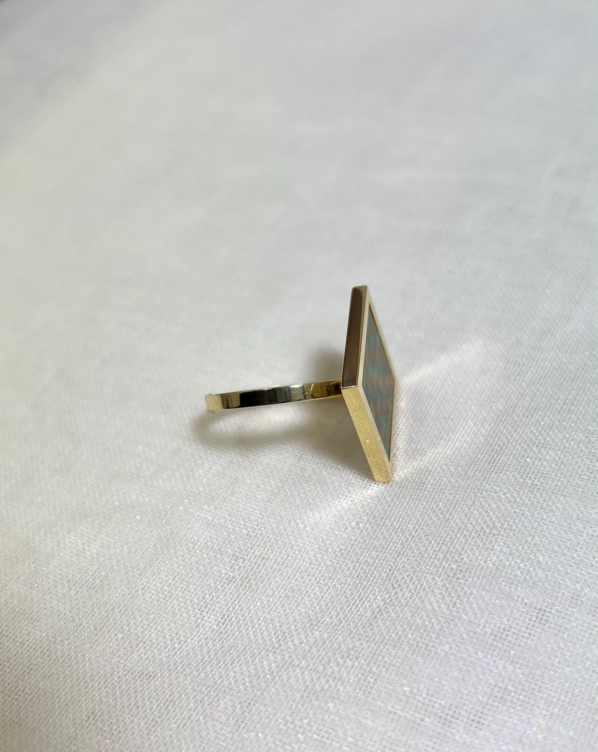 side view of gold square ring with checkerboard design on white fabric background.