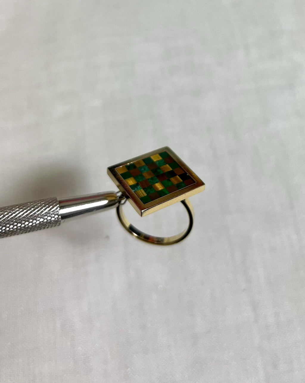 gold square ring with green and brown checkerboard design on white fabric background